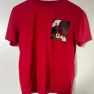 Hugo Boss T-shirt - Röd t-shirt med en cool bröstficka i camouflage-mönster och diskret logga. Klassisk passform och korta ärmar, perfekt för en avslappnad stil.