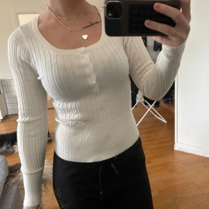Vit ribbad långärmad topp med knappar - Säljer en vit ribbad långärmad topp med rund halsringning och knappdetaljer framtill. Sitter figurnära och har en mjuk, stretchig känsla. Perfekt att styla till både jeans och kjol.