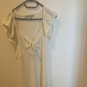 Vit kortklänning med knyt från lager 57 - Söt vit lång klänning från lager 57 med strukturmönster och knyt framtill. Klänningen har v-ringning och volangdetaljer vid axlarna. Perfekt för sommarens varma dagar.