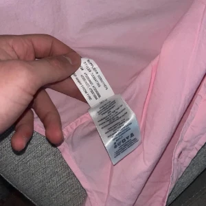 Rosa skjorta från Ralph Lauren - Säljer en klassisk rosa skjorta från Ralph Lauren med blå broderad logga på bröstet. Skjortan har knappar framtill och krage, tillverkad i bomull. Perfekt för dig som gillar stilrena plagg med ikonisk design.