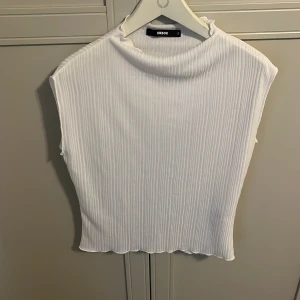 Vit ribbad topp från Bik Bok - Säljer en vit ribbad topp från Bik Bok med hög hals och ärmlös design. Toppen har en lätt croppad passform och är tillverkad i ett stretchigt material som sitter snyggt på kroppen. Perfekt att styla till jeans eller kjol!