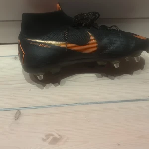 Nike Mercurial fotbollsskor i svart och orange.      (Lägg bud) - Säljer ett par Nike Mercurial fotbollsskor i svart med orange detaljer och snörning. Skorna har högre skaft och är tillverkade i syntetmaterial med tydliga Mercurial-loggor på hälen. Perfekta för dig som vill sticka ut på planen!