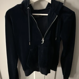 Mörkblå zip hoodie från Juicy Couture - Juicy Couture hoodie i mörkblå velour med dragkedja framtill och huva. Klassisk modell med långa ärmar, ribbade muddar och snörning i huvan. Perfekt för en avslappnad och trendig look.