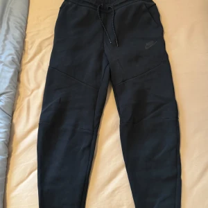 Svarta mjukisbyxor från Nike - Svarta mjukisbyxor från Nike med dragsko i midjan och diskret logga på vänster ben. Byxorna har en normal passform och är tillverkade i mjukt bomullsmaterial. Perfekta för chill eller träning.