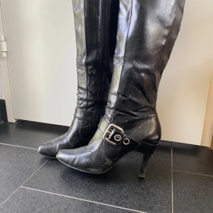 Svarta höga stövlar med spänne och klack - Säljer ett par jätte fina svarta höga stövlar i skinnimitation med  spänne i silver vid ankeln. Stövlarna har smal klack och dragkedja på insidan. 