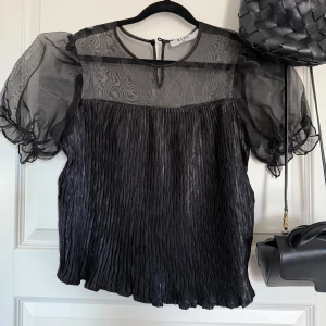 Svart plisserad blus med puffärmar från NA-KD - Säljer en svart plisserad blus från NA-KD med transparenta puffärmar och rund halsringning. Blusen har en lätt glansig finish och knäppning bak i nacken. Perfekt för dig som vill ha en trendig och luftig topp.