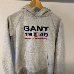 Grå hoodie från GANT - Grå hoodie från GANT med broderad logga och texten '1949 American Sportswear' framtill. Tröjan har huva och magficka, samt långa ärmar. Perfekt för en avslappnad och sportig stil.