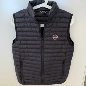 Svart dunväst från Colmar - Snygg svart dunväst från Colmar med dragkedja framtill och två praktiska fickor. Perfekt för lager-på-lager under kyliga dagar. Västen har en stilren design med Colmar-logga på bröstet. Nyskick 