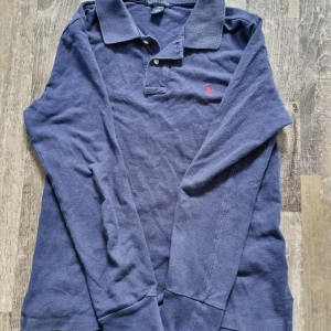 Blå pikétröja från Ralph Lauren - Säljer en klassisk blå pikétröja från Ralph Lauren med lång ärm och en liten röd🤩logotyp på bröstet. Tröjan har en krage och knappar framtill. Perfekt för en stilren look😃