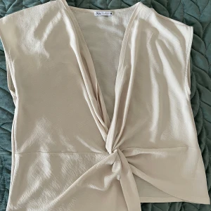 Beige omlottopp från Zara - Säljer en stilren beige omlottopp från Zara. Toppen har en v-ringad design med knytning framtill och är ärmlös. Perfekt för en elegant och avslappnad look.