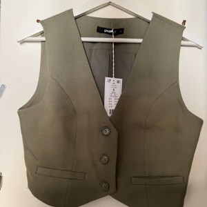 Väst-topp från Gina Tricot - Snygg oanvänd khakifärgad väst från Gina Tricot med knappar framtill. Västen har en klassisk design med två fickor och är tillverkad i ett bekvämt material.❣️