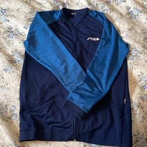 Blå träningsjacka från Stiga - Säljer en snygg blå träningsjacka från Stiga med dragkedja framtill. Jackan har en mörkblå bas med ljusblå ärmar och ett vitt Stiga-tryck på bröstet. Perfekt för sportiga aktiviteter!