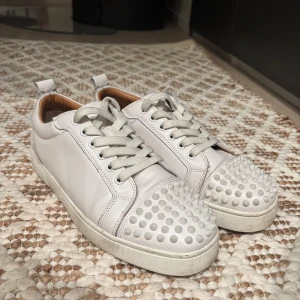 Louboutin - Snygga vita sneakers från Christian Louboutin med coola nitar på tån och klassisk röd sula. Skorna har snörning och är tillverkade i skinn. Perfekta för att ge din outfit en edgy touch.