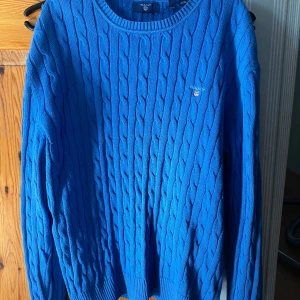 Blå kabelstickad tröja från Gant - Säljer en snygg blå kabelstickad tröja från Gant. Tröjan har en klassisk rund halsringning och ett diskret broderat märke på bröstet. Perfekt för kyligare dagar och ger en stilren look. Passar både till jeans och chinos.