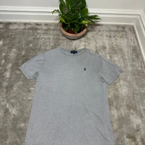 Grå t-shirt från Polo Ralph Lauren - Klassisk grå t-shirt från Polo Ralph Lauren med broderad marinblå logga på bröstet. T-shirten har rund halsringning och korta ärmar. Tillverkad i mjuk bomull för en skön och avslappnad känsla. Perfekt till jeans eller shorts.