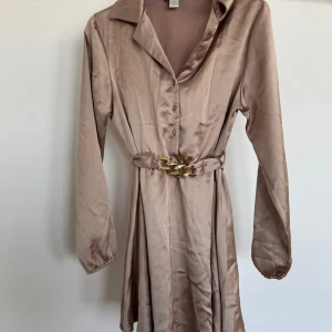 Champagnefärgad sidenklänning med bälte - Elegant champagnefärgad långärmad klänning i siden med knappar framtill och ett dekorativt bälte i midjan. Perfekt för en stilfull look.