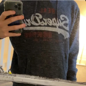 Svart/Grå  hoodie från Superdry - Säljer en snygg mörkblå hoodie från Superdry med tryck framtill. Den har en klassisk passform med långa ärmar och en praktisk ficka på magen. Perfekt för en avslappnad stil. 🚨PRIS KAN DISKUTERAS🚨