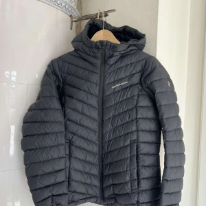 Svart dunjacka från Peak Performance - Snygg och funktionell svart dunjacka från Peak Performance. Jackan har en dragkedja framtill och är quiltad för extra värme. Perfekt för kyliga dagar med sin långa ärm och stilrena design.