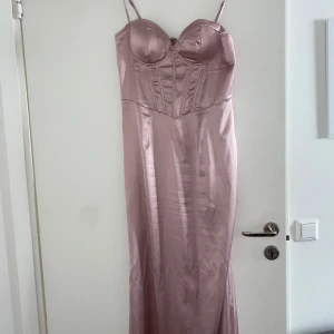 Ljusrosa sidenklänning med smala axelband - Elegant ljusrosa långklänning i siden med smala axelband och en figurnära passform. Klänningen har en korsettliknande överdel som ger en stilren look. Perfekt för speciella tillfällen.