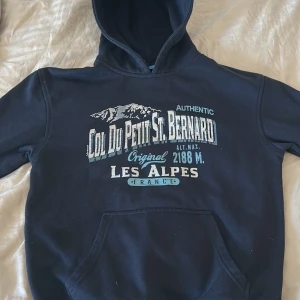 Mörkblå hoodie med tryck - Säljer en mörkblå hoodie med tryck av 'Col du Petit St. Bernard' och 'Les Alpes France' i vitt och ljusblått. Köpt på St. Bernard passet i Frankrike. Väldigt svår att få tag på eftersom den bara säljs på ett ställe. Riktigt fett tryck. Tröjan har en klassisk känguruficka och en bekväm huva. Perfekt för kyliga sommardagar. Nypris: 999kr