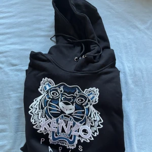 Svart hoodie från Kenzo - Säljer en svart hoodie från Kenzo med en stor broderad tiger på framsidan. Hoodien har en justerbar huva med snörning och är perfekt för en avslappnad stil. Passar bra till både jeans och joggers.