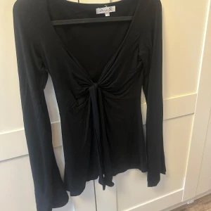 Svart långärmad topp från Design by Us - Snygg svart långärmad topp från Design by Us med knytning framtill. Perfekt för en stilren look. Toppen har en v-ringad design och är tillverkad i ett mjukt material.