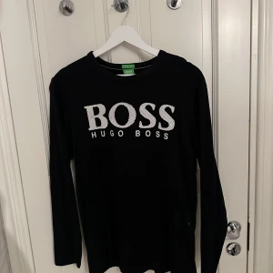 Svart långärmad tröja från Hugo Boss - Snygg svart långärmad tröja från Hugo Boss med vit logga på bröstet. Perfekt för en stilren look. Tröjan har en klassisk passform och är tillverkad i ett mjukt material för extra komfort.