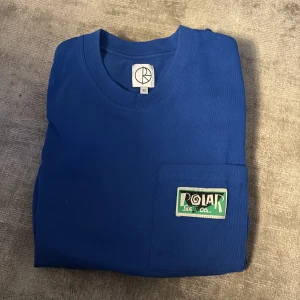 Blå t-shirt från Polar Skate Co. - Snygg blå t-shirt från Polar Skate Co. med en bröstficka och ett coolt märke på framsidan. Perfekt för en avslappnad stil. Rätt baggy