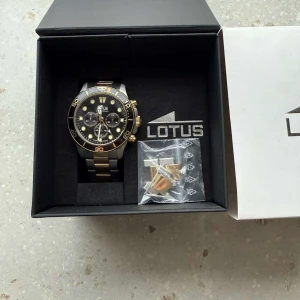 Lotus Chronograph herrklocka - Snygg Lotus Chronograph herrklocka med en kombination av silver och guld på armbandet. Klockan har en svart urtavla med guldiga detaljer och flera sub-dials. Perfekt för den som vill ha en stilren och funktionell accessoar.