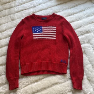 Röd stickad tröja från Polo Ralph Lauren - Säljer en snygg röd stickad tröja från Polo Ralph Lauren med en amerikansk flagga på framsidan. Tröjan har långa ärmar och ribbade muddar vid ärmslut och nederkant. Perfekt för en stilren look!