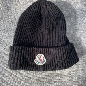 Svart ribbad mössa från Moncler - Snygg svart ribbad mössa från Moncler med deras ikoniska logga framtill. Perfekt för att hålla värmen under kyliga dagar. Enkel och stilren design som passar till det mesta. Ej äkta