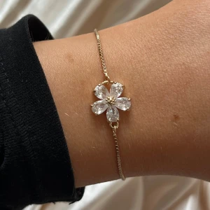 Guldarmband med blomma - Elegant guldfärgat armband med en vacker blomma dekorerad med klara stenar. Armbandet har en justerbar kedja med två små kulor i ändarna för perfekt passform. (Vet inte om den är vattentät eller inte, undvik kontakt med vatten) PRISET GÅR ATT PRUTA!