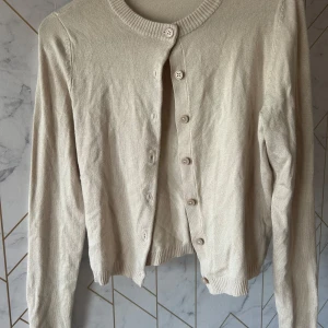 Beige kofta med knappar - Säljer en stilren beige kofta med knappar framtill. Den har långa ärmar och en rund halsringning. Perfekt för lager på lager-stil och passar till många olika outfits. Hör av dig vid intresse!