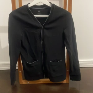 Svart cardigan - Säljer en stilren svart cardigan från WE med knappar framtill och två fickor. Perfekt för en elegant och avslappnad look. Koftan har långa ärmar och en enkel design som passar till många tillfällen. Hör av er vid frågor!