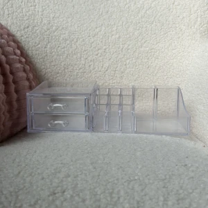 Transparent sminkförvaring med lådor - Praktisk och stilren transparent sminkförvaring med två lådor och flera fack för organisering av sminkprodukter. Perfekt för att hålla ordning på sminkborstar, läppstift och andra små tillbehör.