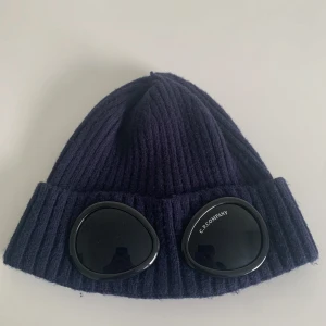 Mörkblå mössa med glasögondetalj från C.P. Company - Hej! Säljer nu min Snygga mörkblå mössa från C.P. Company med unika glasögondetaljer framtill. Den är ribbstickad och ger en cool look. Perfekt för att hålla värmen med stil. Den är köpt för 900kr och är använd ett par gånger under hösten. Mössan har inga stora defekter, den är sydd på toppen. Priset är inte fast det kan även diskuteras vid snabb affär, om du undrar något så hör av dig😄