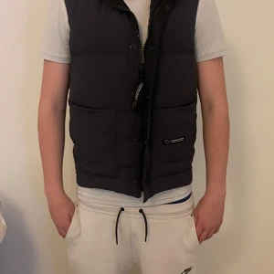Svart dunväst från Canada Goose - Säljer en svart dunväst från Canada Goose. Västen har en dragkedja och knappar framtill samt två stora fickor. Perfekt för kyligare dagar. Inget og ingår köpt för 7999kr på NK i Stockholm. Skriv om ni undrar något!