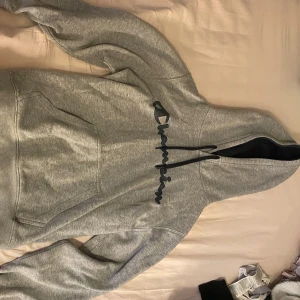 Grå hoodie från Champion - Säljer en grå hoodie från Champion med svart logga på bröstet. Tröjan har en klassisk design med känguruficka och justerbar huva. Perfekt för en avslappnad stil.