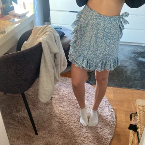 Blå blommig kjol med volang - Söt blå kjol med blommigt mönster och volangdetaljer. Kjolen har en omlott-design och knytning i midjan, vilket ger en fin passform. Perfekt för sommarens varma dagar!