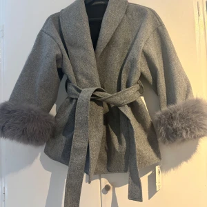 Grå kappa med pälsdetaljer - Snygg grå kappa med knytbälte och fluffiga pälsdetaljer vid ärmsluten. Perfekt för kyliga dagar när du vill vara både varm och stilren. Den har en elegant omlott-design som ger en chic look.