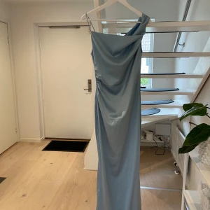 Ljusblå långklänning från ADOORE - Elegant ljusblå långklänning från ADOORE med en stilren oneshoulder-design.       Modell Almeria i light blue. Perfekt för speciella tillfällen med sin smickrande passform och mjuka material.             97% polyester, 3% elastane                          Använd vid ett tillfälle                   OBS! Bild från hemsida visar likadan klänning fast i grönt. Den jag säljer är ljusblå 