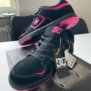 Svarta och rosa sneakers från DC - Snygga svarta sneakers från DC med rosa detaljer och logga. Skorna har en robust sula med mönster för bra grepp och snörning för justerbar passform. Perfekta för en cool och avslappnad stil. Helt nya med tag 