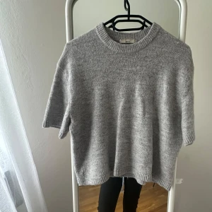 Grå stickad t-shirt från Gina Tricot - Säljer en stilren grå stickad tröja från Gina Tricot. Tröjan har korta ärmar och en rund halsringning, perfekt för lager-på-lager eller som den är. Super fin men säljer då den tyvärr inte kommer till användning längre.