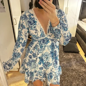 Blommig klänning med spetsdetaljer - Säljer en söt blommig klänning i vitt och blått med v-ringad spetsdetalj. Klänningen har långa ärmar och en volangkant nedtill. Perfekt för en somrig look! Lite tajt på mig som har s- m så skulle säga att de passar bättre på nån som har xs-s
