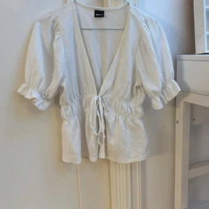 Vit blus från Gina Tricot - Charmig vit blus från Gina Tricot med knytning framtill och elastisk midja. Blusen har korta puffärmar och en v-ringad design som ger en romantisk touch. Perfekt för en somrig look!