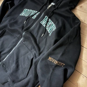 Svart zip hoodie med grön text - Säljer en svart hoodie med texten 'MOVEMENT' i vitt och grönt på bröstet. Hoodien har dragkedja och fickor framtill samt en huva med snören. Perfekt för en avslappnad stil.