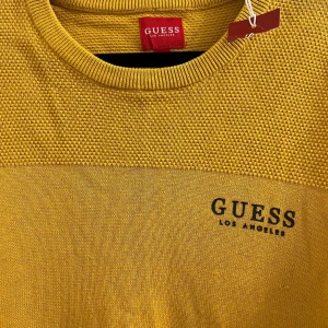 Gul tröja från Guess - Säljer en snygg gul tröja från Guess med texten 'Guess Los Angeles' tryckt på bröstet. Tröjan har ett strukturerat mönster i tyget och långa ärmar. Perfekt för en stilren look.