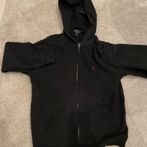 Svart hoodie från Polo Ralph Lauren - Säljer en klassisk svart hoodie från Polo Ralph Lauren med dragkedja och huva. Den har en liten röd logga på bröstet och praktiska fickor framtill. Perfekt för en avslappnad stil. Axelbredd: 41 cm längd: 60 cm