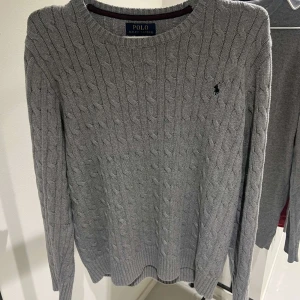 Grå stickad tröja från Ralph Lauren - Säljer en stilren grå stickad tröja från Ralph Lauren med kabelstickat mönster. Tröjan har långa ärmar och en liten broderad logotyp på bröstet. Perfekt för kyligare dagar!