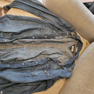 Blå jeansskjorta från Hilfiger Denim - Snygg blå jeansskjorta från Hilfiger Denim med klassisk design och knappar framtill. Skjortan har långa ärmar och två bröstfickor med knappstängning. Perfekt för en avslappnad stil.